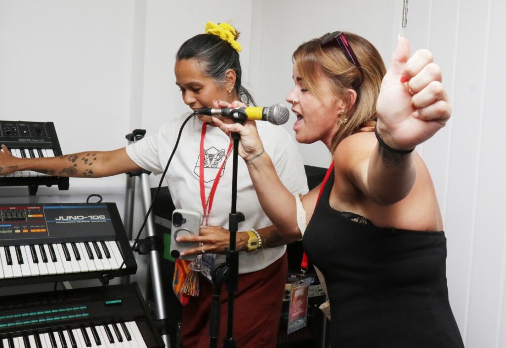A person singing into a microphone with some keyboards in the background