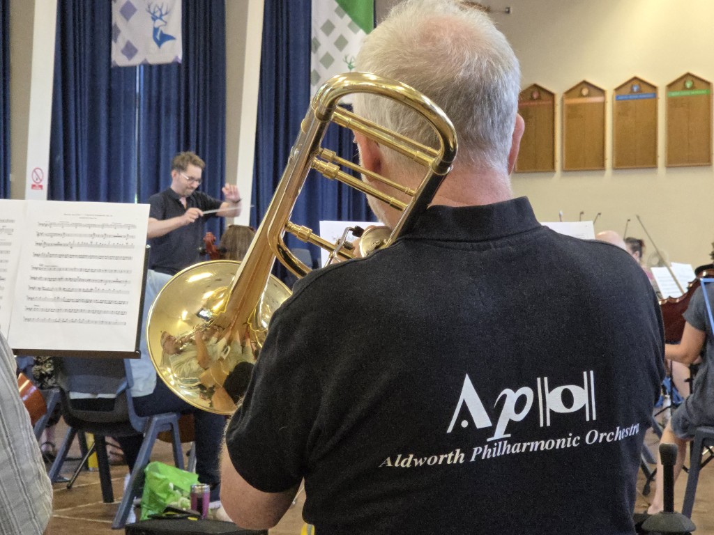 An over-the-shoulder view of a male trombonist from behind, wearing a black polo shirt with the white Aldworth Philharmonic Orchestra logo on the back. He holds a brass trombone, with the reflective bell of the instrument visible. In the blurred background, musical director Andrew Taylor stands on a podium conducting the orchestra.
