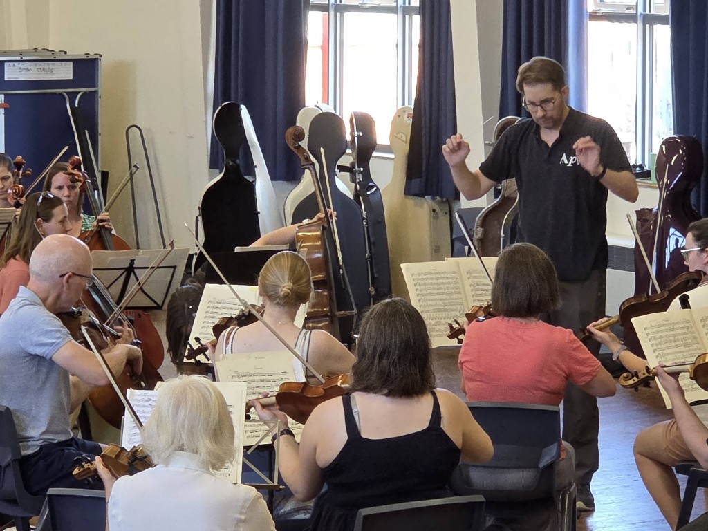 Aldworth Philharmonic Orchestra musical director Andrew Taylor stands and conducts a group of musicians playing string instruments, including violins and cellos, in a rehearsal room. The musicians are seated with sheet music on stands in front of them, while empty cello cases stand against the wall behind them.
