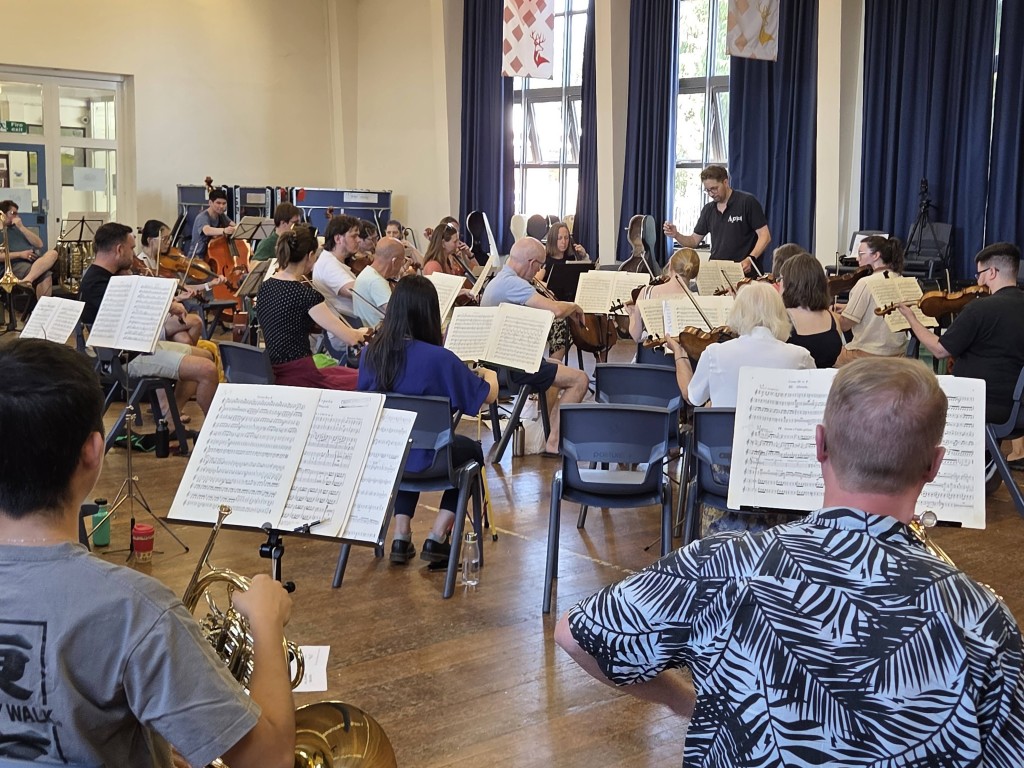 A wide-angle shot of the Aldworth Philharmonic Orchestra rehearsing in a large hall with wooden flooring and high windows. Musicians playing brass, woodwind, and string instruments are seated in semi-circles with their sheet music stands. Musical director Andrew Taylor is visible in the background, conducting the full ensemble.