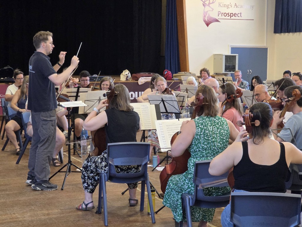 Musical director Andrew Taylor stands on the left holding a conducting baton, guiding the Aldworth Philharmonic Orchestra. Seated cello and violin players face toward him in a semi-circle. A sign in the background reads King's Academy Prospect.
