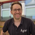 A close-up headshot of Andrew Taylor, the musical director of the Aldworth Philharmonic Orchestra. He is smiling warmly at the camera, wearing glasses and a black polo shirt featuring a white orchestra logo. The background is a softly blurred outdoor school building setting.