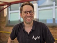 A close-up headshot of Andrew Taylor, the musical director of the Aldworth Philharmonic Orchestra. He is smiling warmly at the camera, wearing glasses and a black polo shirt featuring a white orchestra logo. The background is a softly blurred outdoor school building setting.