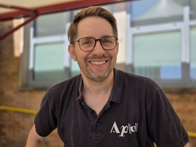 A close-up headshot of Andrew Taylor, the musical director of the Aldworth Philharmonic Orchestra. He is smiling warmly at the camera, wearing glasses and a black polo shirt featuring a white orchestra logo. The background is a softly blurred outdoor school building setting.