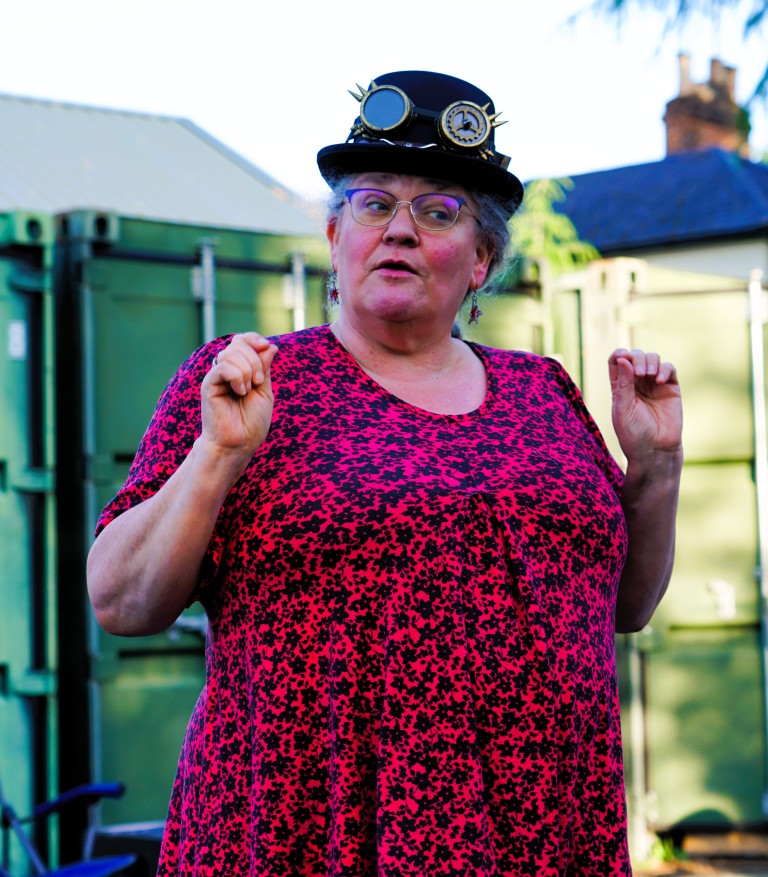 A woman wearing a streampunk style hat, holding her arms aloft; they are bent at the elbow. She is tilting her head, and wearing a red and black patterned dress. 