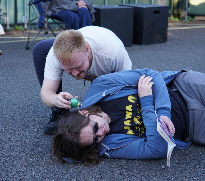 A rehearsal image of Hamlet. A man is lying on the ground and a second man is appearing to pour something into his ear