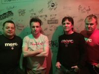 A colour photograph of the four male members of the band The Chords UK standing in a line from left to right against a light-coloured wall covered in band signatures and graffiti. They are all looking towards the camera with serious expressions. Three of the members are wearing t-shirts or long-sleeved tops from the brand Merc London, featuring a Union Jack design and a mod target logo. The lighting has a strong red and green cast across the scene.