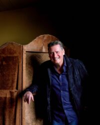 Tony Hadley wearing a blue shirt and jacket leaning against a brown dresser