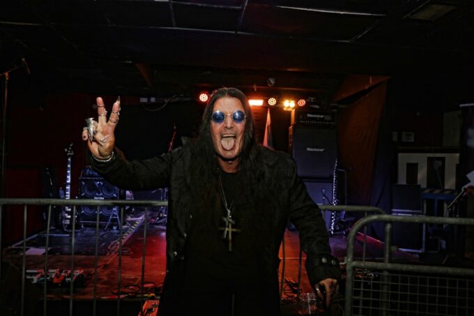 Sabbra Cadabra vocalist Kevin McGillian standing on stage in character as Ozzy Osbourne, wearing round blue sunglasses, a long dark coat, and a large cross pendant while making a peace sign at the Face Bar in Reading