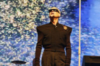Philip Oakey looking upwards past a white microphone stand wearing a black structured coat with a brooch against a colourful digital screen backdrop