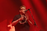 Craig Reid of The Proclaimers wearing glasses and singing into a microphone on stage while holding up a white tambourine against red stage lighting