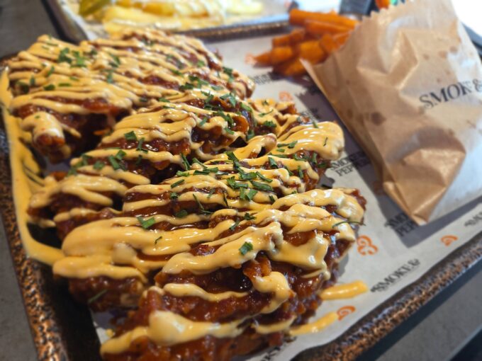 A photograph taken from above and to the side, looking down upon a metal tray containing a portion of dark, sauce-smothered chicken tenders and a cluster of orange sweet potato fries. An brown paper bag with the black text "SMOKE & PEPPER" lies in the foreground. Another dish of regular fries is partially visible in the blurry background.