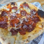 An overhead close-up shot of a sourdough pizza served on branded greaseproof paper on a wooden bench. The pizza displays a golden-brown crust with charred bubbles, topped with rich red tomato sauce, melted cheese, pepperoni slices, sliced red onions, and finely grated cheese.