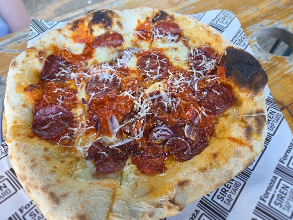 An overhead close-up shot of a sourdough pizza served on branded greaseproof paper on a wooden bench. The pizza displays a golden-brown crust with charred bubbles, topped with rich red tomato sauce, melted cheese, pepperoni slices, sliced red onions, and finely grated cheese.