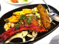 A sizzling iron plate filled with tandoori lamb chops, chicken tikka, a sheek kebab, and sliced onions garnished with fresh coriander