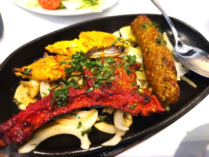 A sizzling iron plate filled with tandoori lamb chops, chicken tikka, a sheek kebab, and sliced onions garnished with fresh coriander