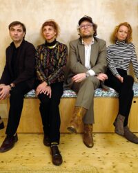 Alt text Four members of the indie rock band Aircooled sit together on a wooden bench against a textured wall. From left to right, Justin Welch wears a dark jacket, Riz Maslen wears a colourful patterned top with black trousers, Oliver Cherer wears a grey suit, glasses, cap, and brown cowboy boots, and Katharine Wallinger wears a black-and-white striped roll-neck top and ankle boots.