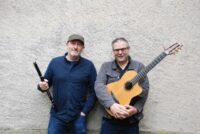 Celtic folk musicians Michael McGoldrick holding a black wooden flute and Tim Edey holding an acoustic guitar while standing against a textured stone wall.