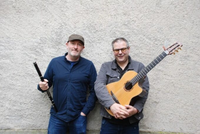 Celtic folk musicians Michael McGoldrick holding a black wooden flute and Tim Edey holding an acoustic guitar while standing against a textured stone wall.