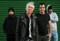 The four members of punk band TV Smith & The Bored Teenagers standing together outdoors against a green corrugated metal shutter door. TV Smith stands in the centre wearing a black leather jacket over a dark shirt with a torn tie, flanked by his three bandmates dressed in casual hoodies and t-shirts