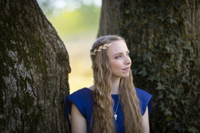 Organist Anna Lapwood with long wavy hair wearing an Elven style gold circlet headpiece and blue dress, leaning between two tree trunks outdoors
