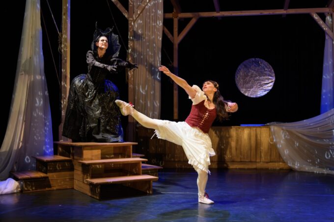 A dancer in a white dress balances on one leg while a performer dressed as the Queen of the Night reaches forward from wooden steps