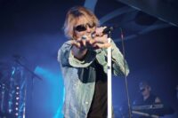 Lead vocalist of tribute band Bon Giovi singing into a microphone on a white stand while reaching his hand towards the crowd under blue stage lights, wearing sunglasses and a denim jacket.