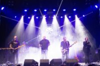 Dark Side of the Wall performing live on stage surrounded by theatrical smoke, illuminated by white spotlights with a round logo projected behind them