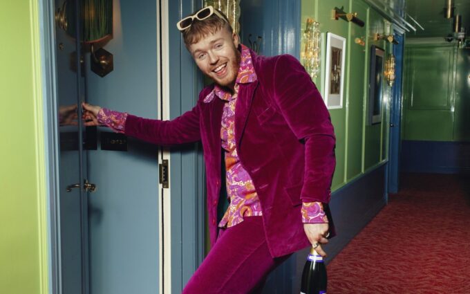 Comedian Finlay Christie smiling in a purple velvet suit, patterned shirt, and sunglasses on his head while holding a bottle of champagne in a hotel corridor.