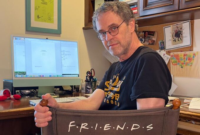 American television writer and showrunner Greg Malins wearing round spectacles and leaning over a director's chair printed with the Friends logo in an office setting