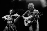 Black and white photograph of Grumpy O Sheep performing on stage under stage lighting, with a woman playing violin on the left and a man playing acoustic guitar on the right