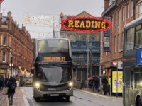 A modern Reading Buses double-decker bus travelling along a town centre street beneath an illuminated Welcome to Reading sign