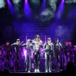 Tribute artist Ben Bowman performs as Michael Jackson in a silver and black jacket flanked by dancers in black leather outfits under purple stage lights and theatrical haze