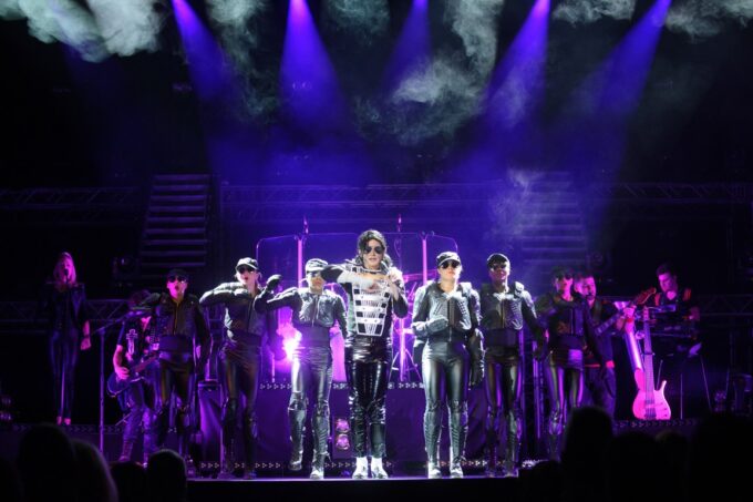 Tribute artist Ben Bowman performs as Michael Jackson in a silver and black jacket flanked by dancers in black leather outfits under purple stage lights and theatrical haze