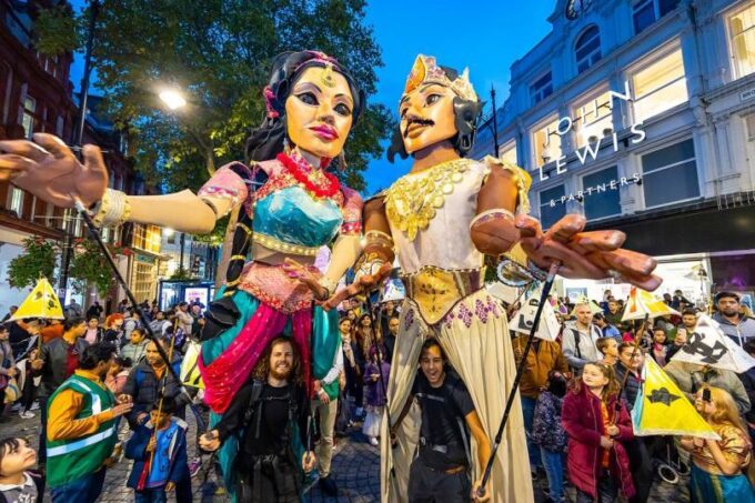 Two giant puppets dressed in colourful traditional South Asian attire being operated by performers amongst crowds carrying paper lanterns outside John Lewis & Partners on Broad Street at dusk
