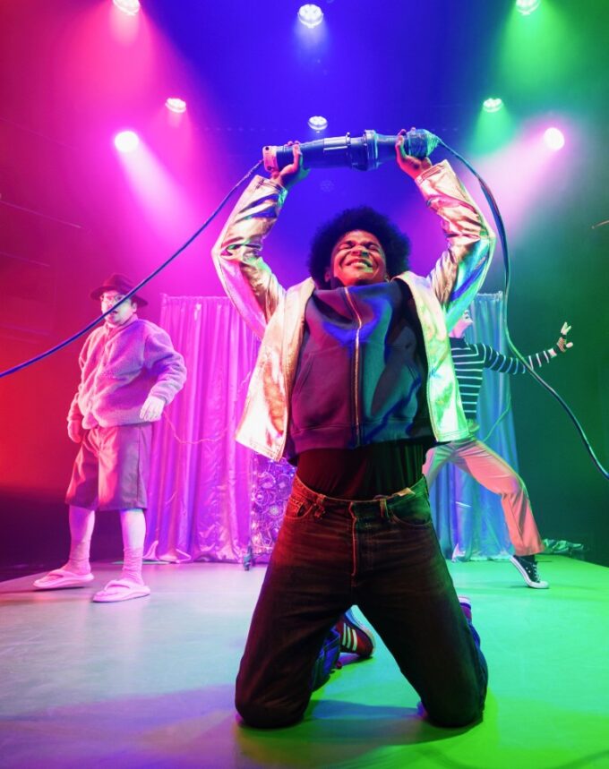 Three performers in The Shivers on a blue-lit stage, one in a gold jacket pointing skywards while two others crouch on all fours
